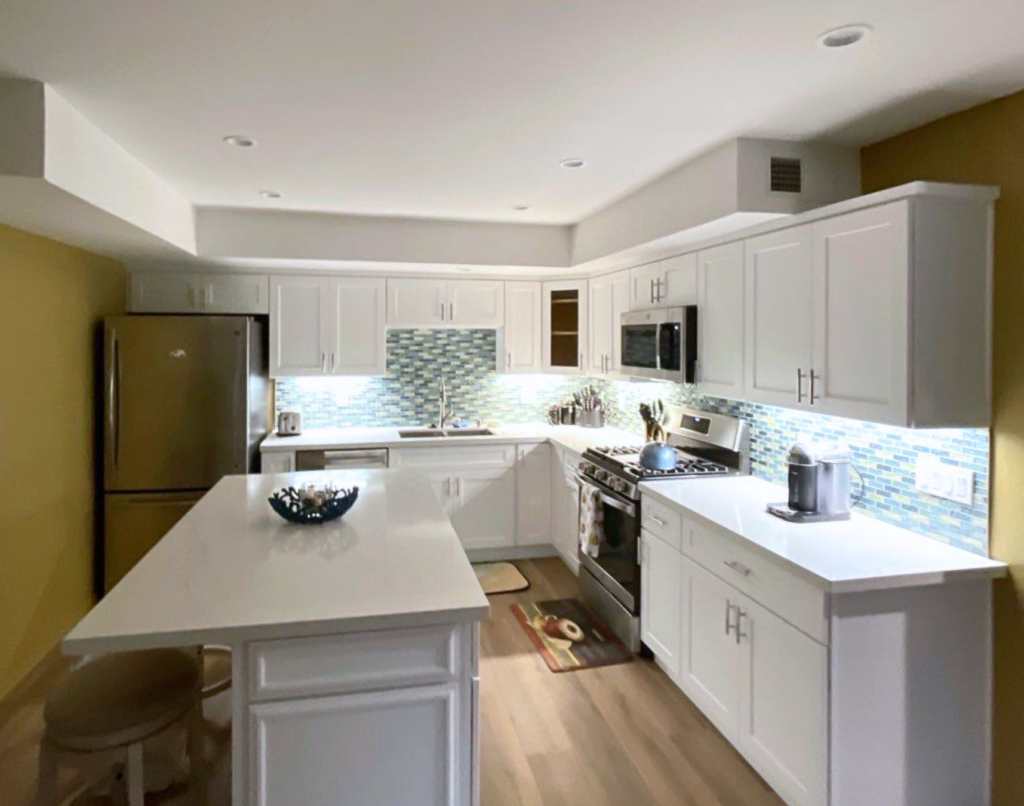 Modern kitchen interior with white cabinetry, stainless steel appliances, a central island, and a multicolored tile backsplash.