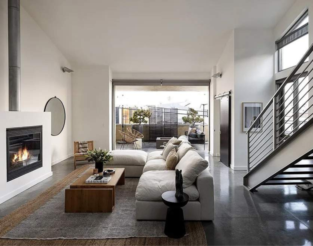 A modern living room with a lit fireplace, a sectional sofa, a wooden coffee table on a gray area rug, a steel staircase leading to an upper level, and sliding glass doors opening to an outdoor area.