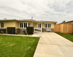A single-story house with a beige exterior, a white fence, and a wooden gate on a cloudy day. The house has a manicured lawn, a concrete pathway leading to the front door, and shrubbery flanking the entrance.