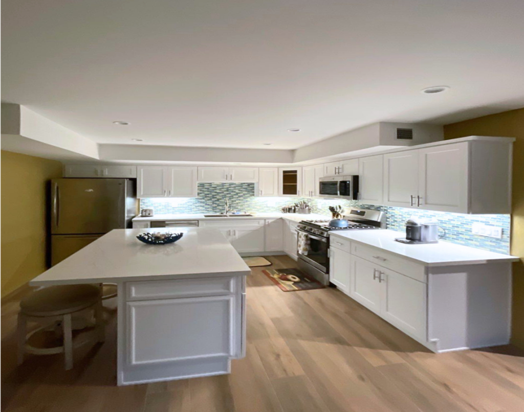 A modern kitchen interior with white cabinetry, stainless steel appliances, and a central island with a white countertop. The backsplash features blue and gray tiles, and there is light wood flooring throughout. A bowl is placed on the island and a small mat lies in front of the stove.