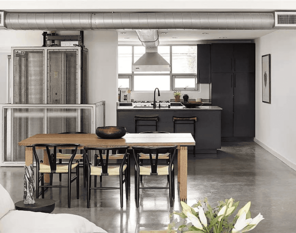 A modern industrial kitchen with stainless steel appliances, exposed ductwork, a polished concrete floor, a wooden dining table with black chairs, and dark cabinetry. Natural light streams in from a window over the sink.