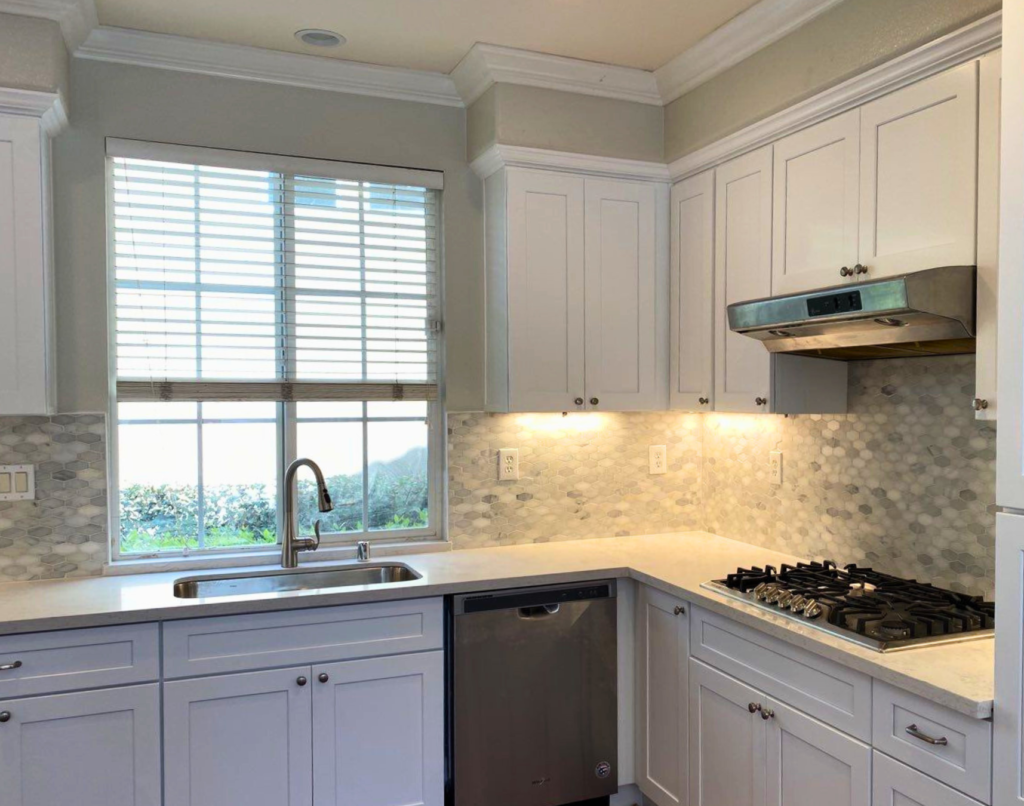 A modern kitchen with white cabinetry and stainless steel appliances, featuring a gas stove, range hood, and dishwasher. The backsplash is tiled with hexagonal patterns, and there's a sink with a view of nature through a window covered with horizontal blinds.