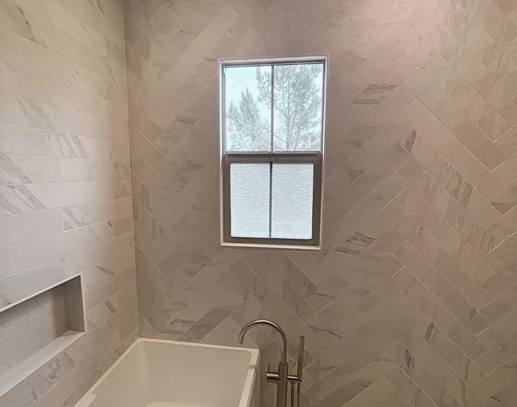 A modern bathroom with herringbone-patterned tile walls, a built-in bathtub, and a window showing trees and a lake outside.