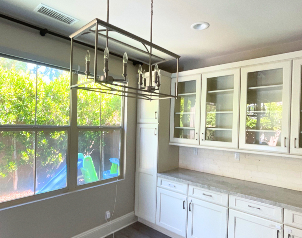 A modern kitchen interior with white cabinetry, a subway tile backsplash, and a large window overlooking greenery outside. In the foreground, an industrial-style chandelier hangs over the space.