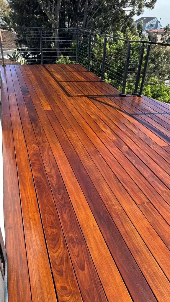Newly installed hardwood flooring on a balcony with a modern railing, showcasing rich wood grain and a polished finish, surrounded by greenery and residential rooftops.